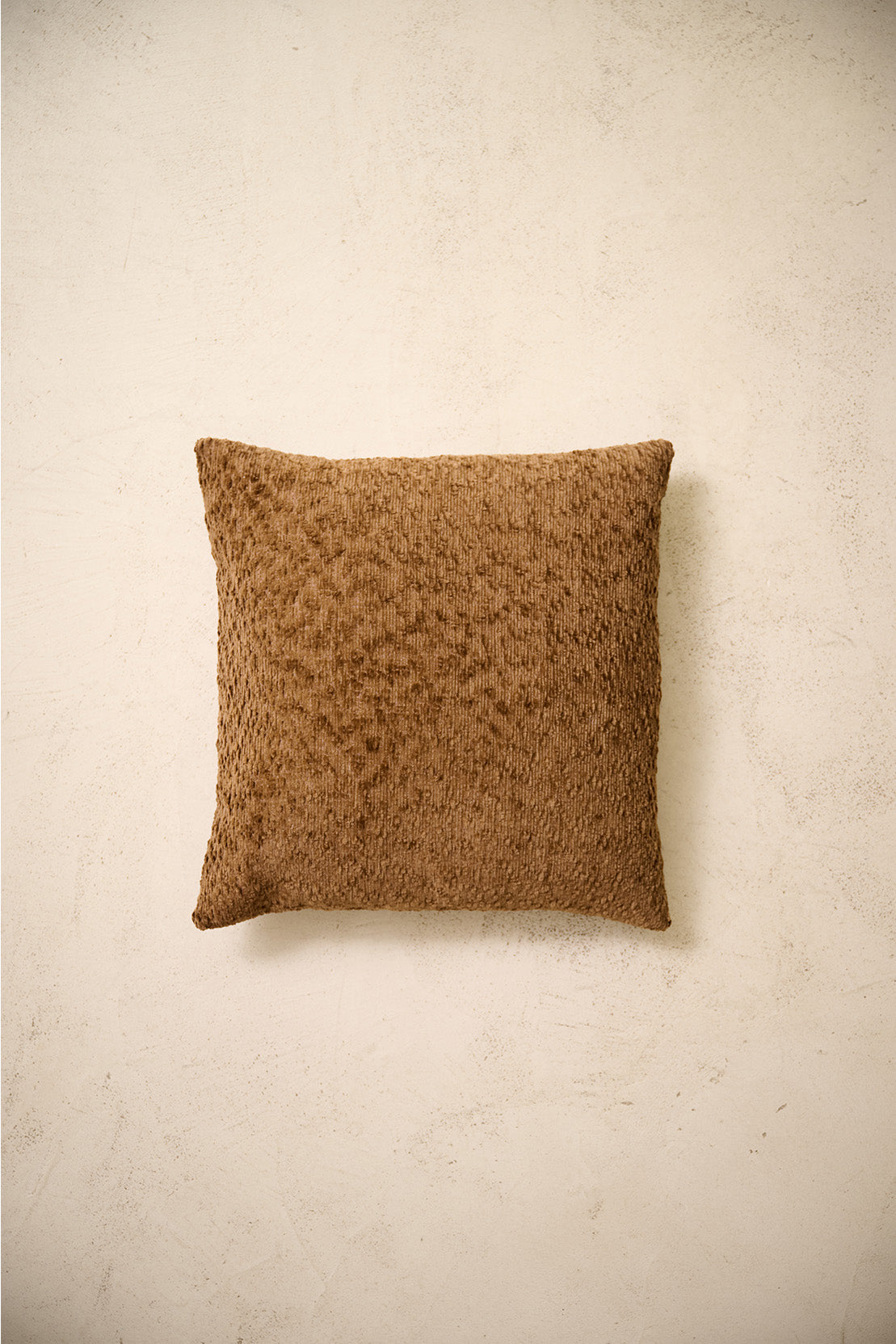 coussin carré CASTLE au relief bouclé brun caramel, posé sur un mur en enduit crème