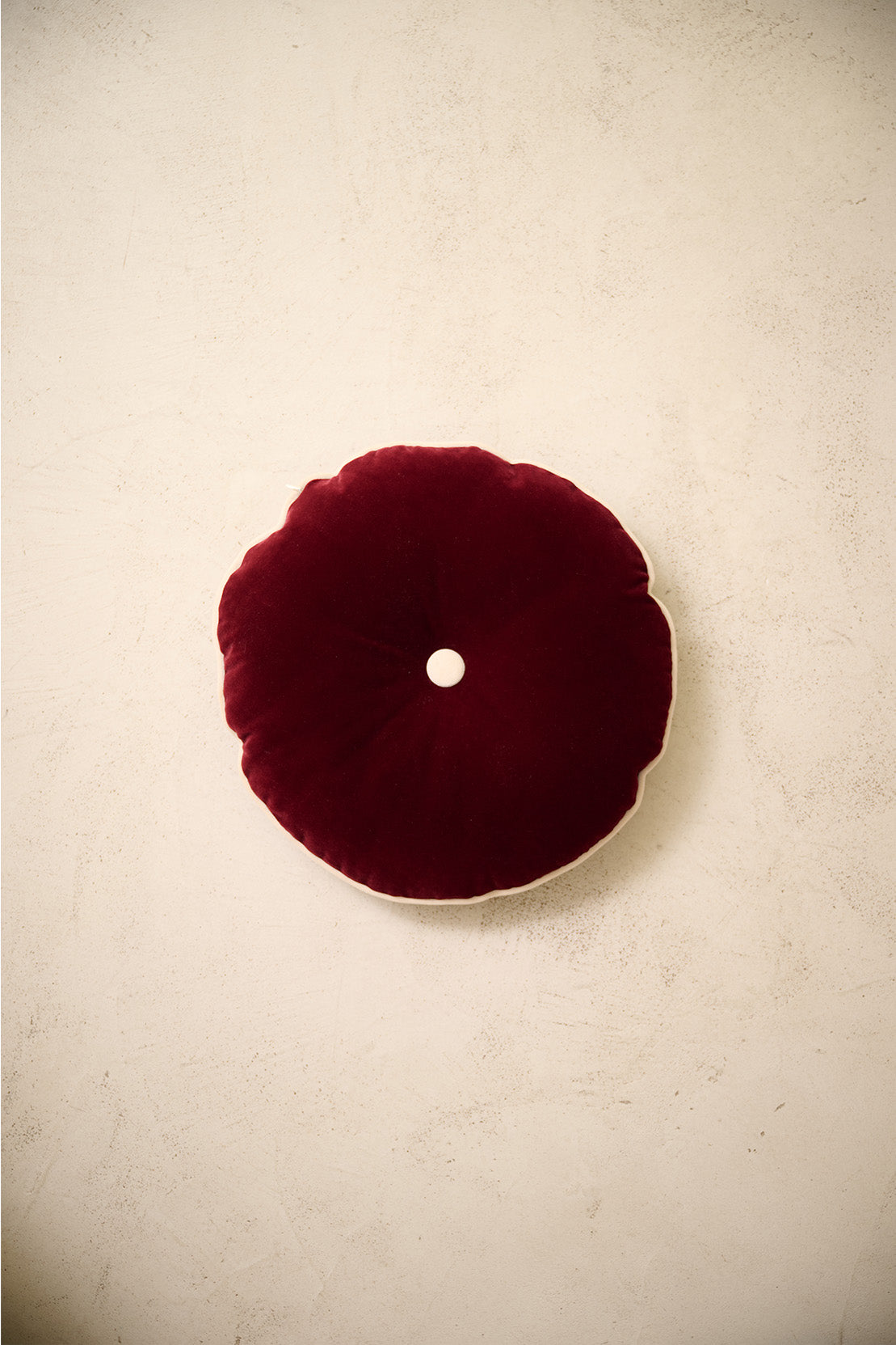 coussin rond MOON en velours rouge framboise à bouton écru, sur un mur en enduit crème