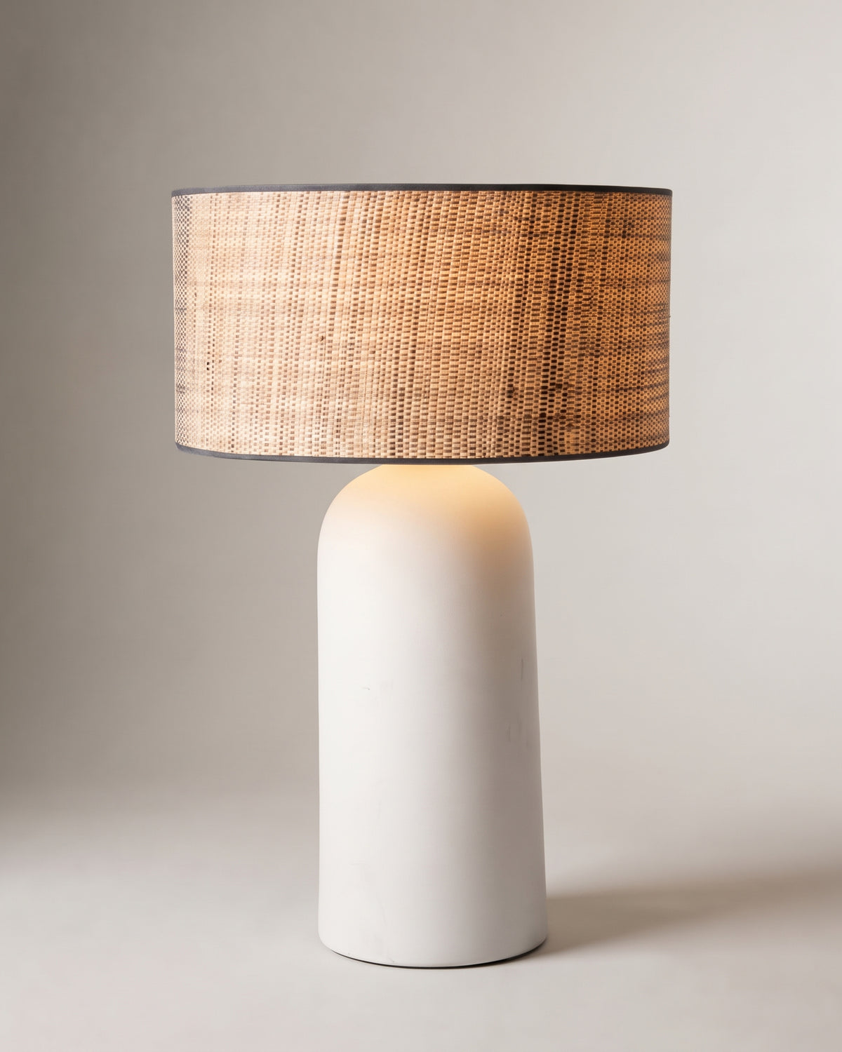 lampe de table AGAPE allumée, pied en céramique blanche mate et abat-jour en rotin tressé