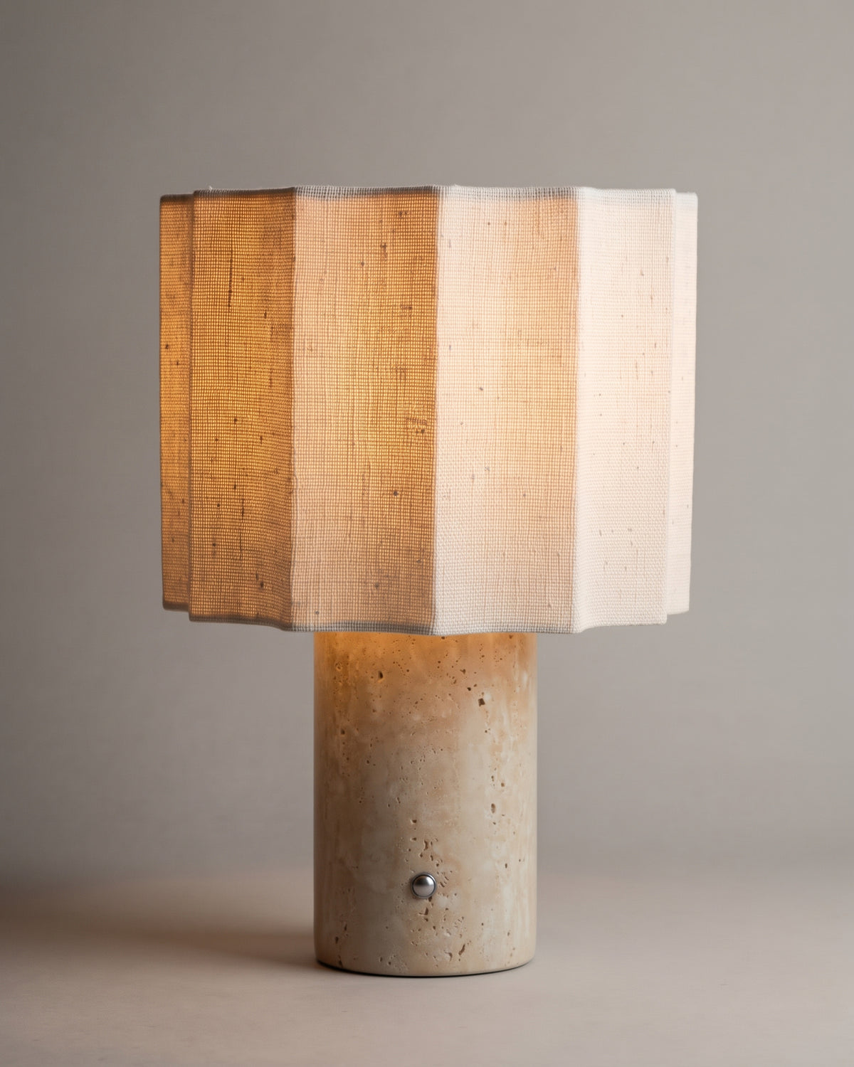 lampe de table BRIDGET allumée, pied cylindrique en travertin et abat-jour en lin à pans