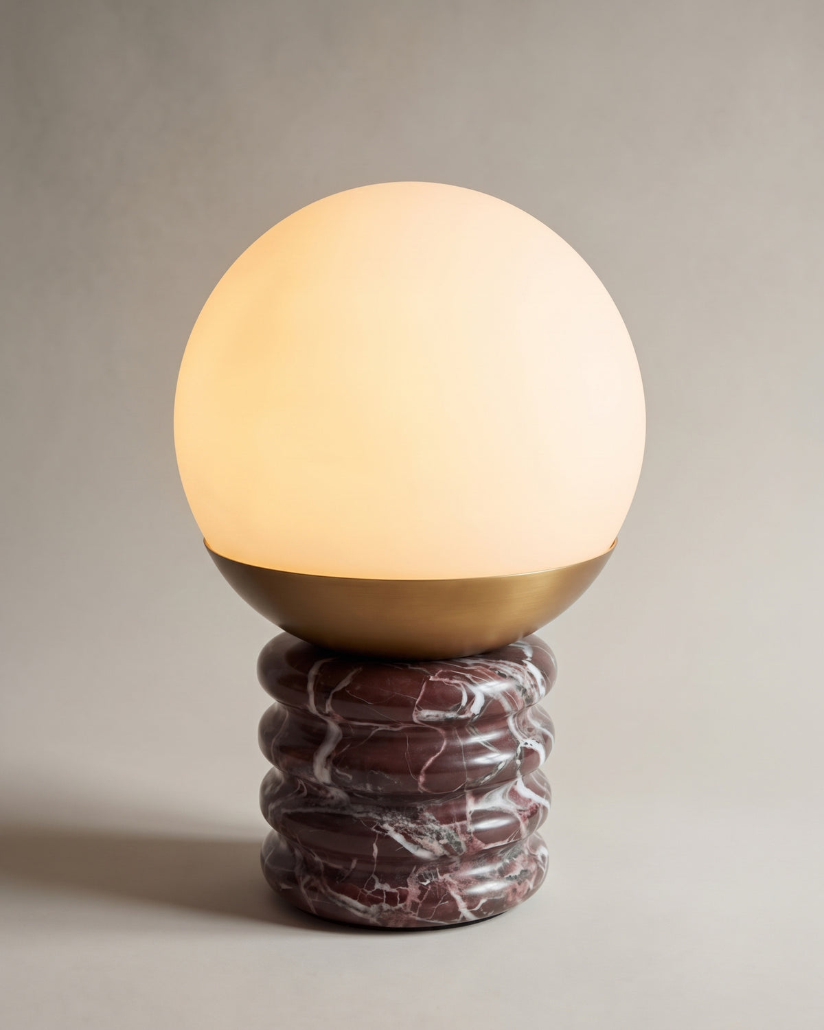 lampe de table NOCTIS allumée, globe opalin et laiton sur un socle ondulé en marbre rouge